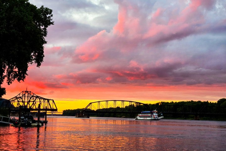 Sunset over river with bridge silhouette and boat, colorful sky with pink and orange clouds.