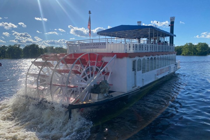 Paddlewheel riverboat on water with sunny sky and trees in background.